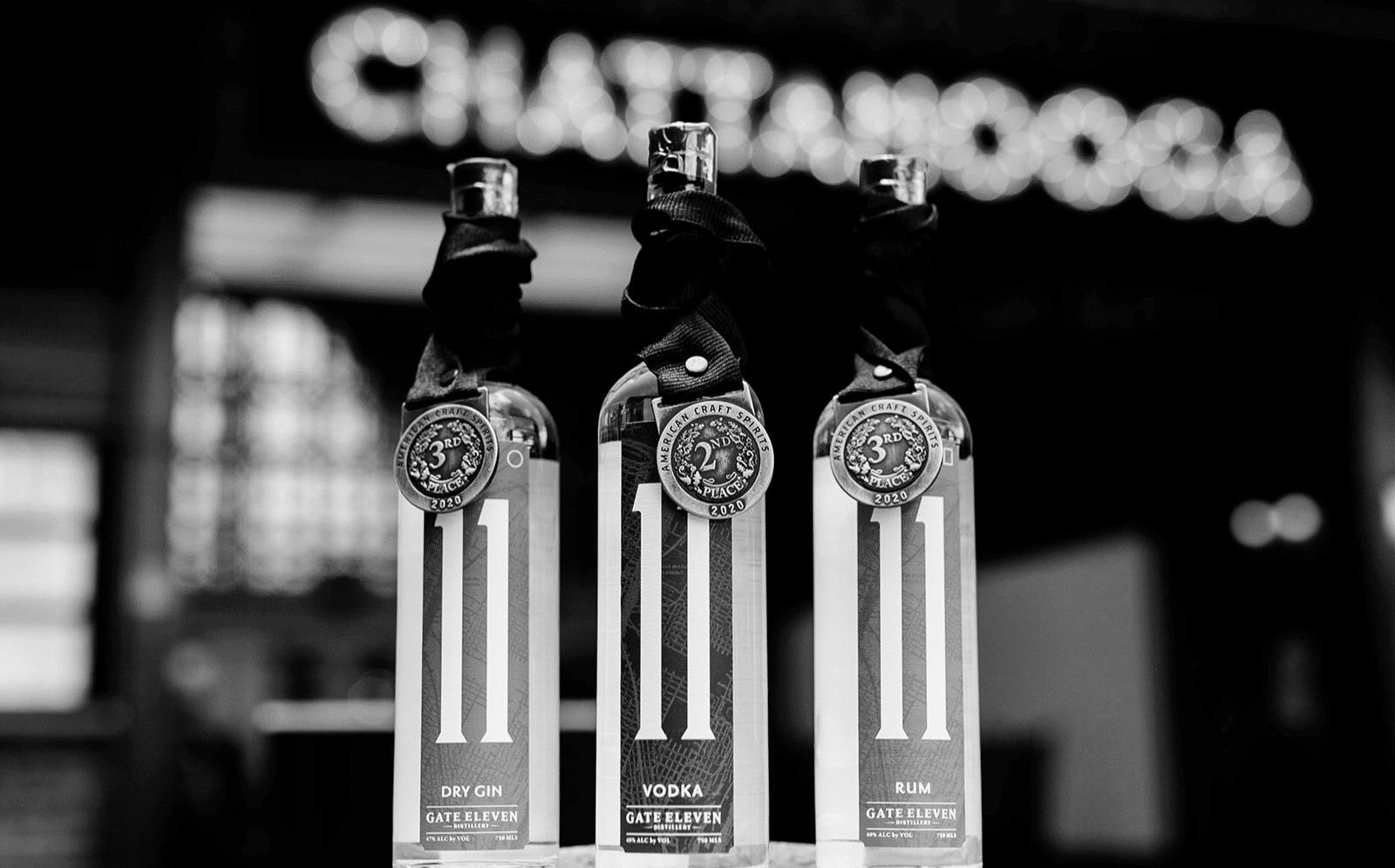 Black and white image of three liquor bottles labeled Gate Eleven. The bottles are arranged side by side, each with a different type: Dry Gin, Vodka, and Rum. "CHATTANOOGA" is blurred in the background.