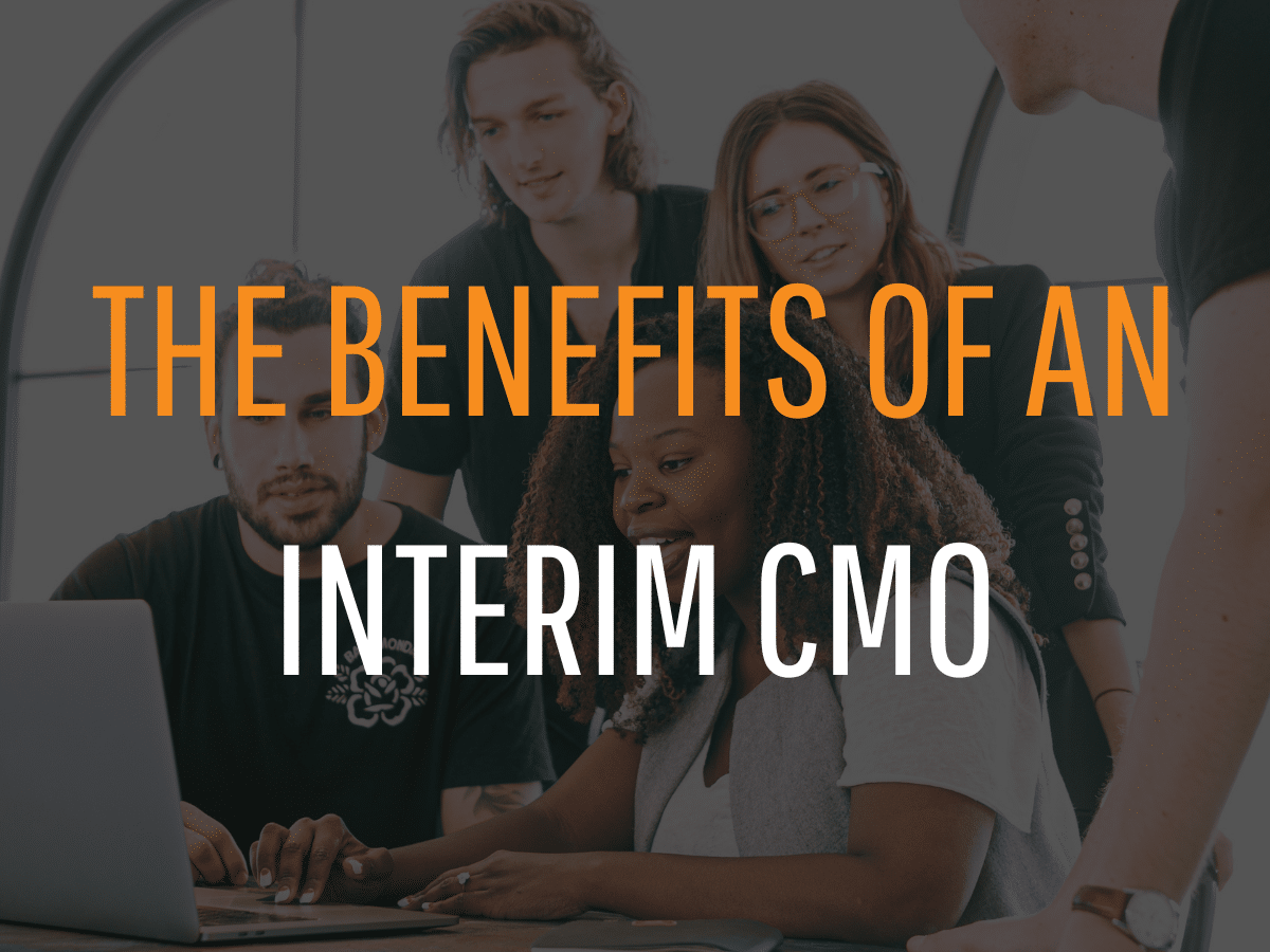 A diverse group of five people is gathered around a table, looking at a laptop screen. Text overlay reads, "The Benefits of an Interim CMO for Your Marketing Strategy." The setting appears to be a modern office space with large windows in the background.