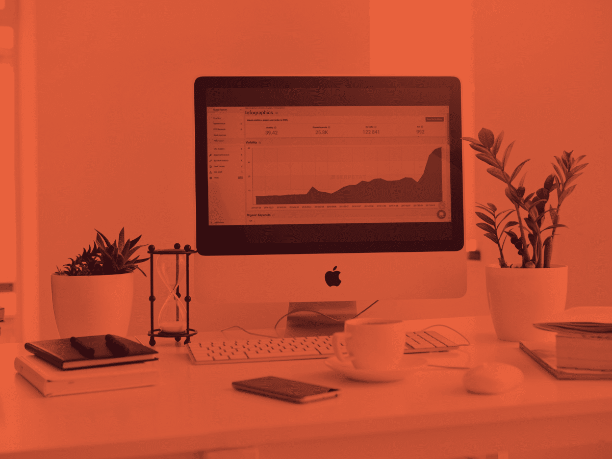 A desktop computer displaying a graph is placed on a white desk. The desk also has potted plants, a cup, a notebook, a smartphone with Generate Responsive Search Ads open, and an hourglass. The image is tinted with an orange hue.