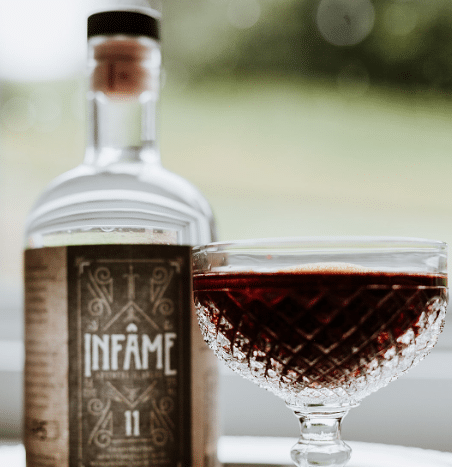 A close-up of a vintage-style glass filled with a red liquid, positioned in front of a bottle labeled "INFAME 11." The glass, with intricate etchings, exemplifies pursuing perfection, and the rustic design of the bottle complements it. The background is softly blurred, focusing attention on the glass and bottle.