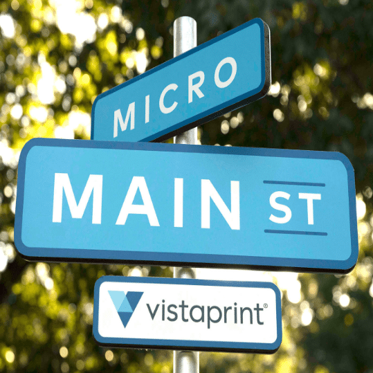 A street sign with blue panels showing "MICRO" and "MAIN ST" intersecting, with a "vistaprint" logo on a separate small rectangular sign below, promoting their printing services. Background features blurred green and yellow foliage.