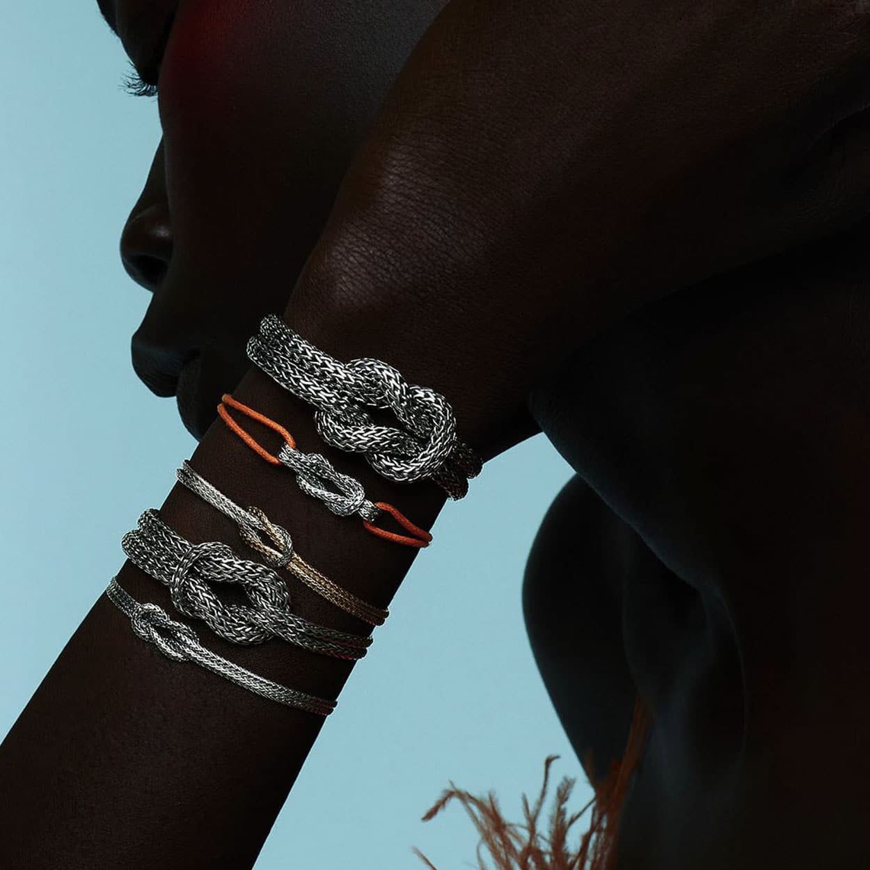 Close-up of an individual showcasing multiple intricately designed silver bracelets with a knot motif on their forearm against a light blue background. Some bracelets have orange accents, and the person's face is partially visible, turned to the side.
