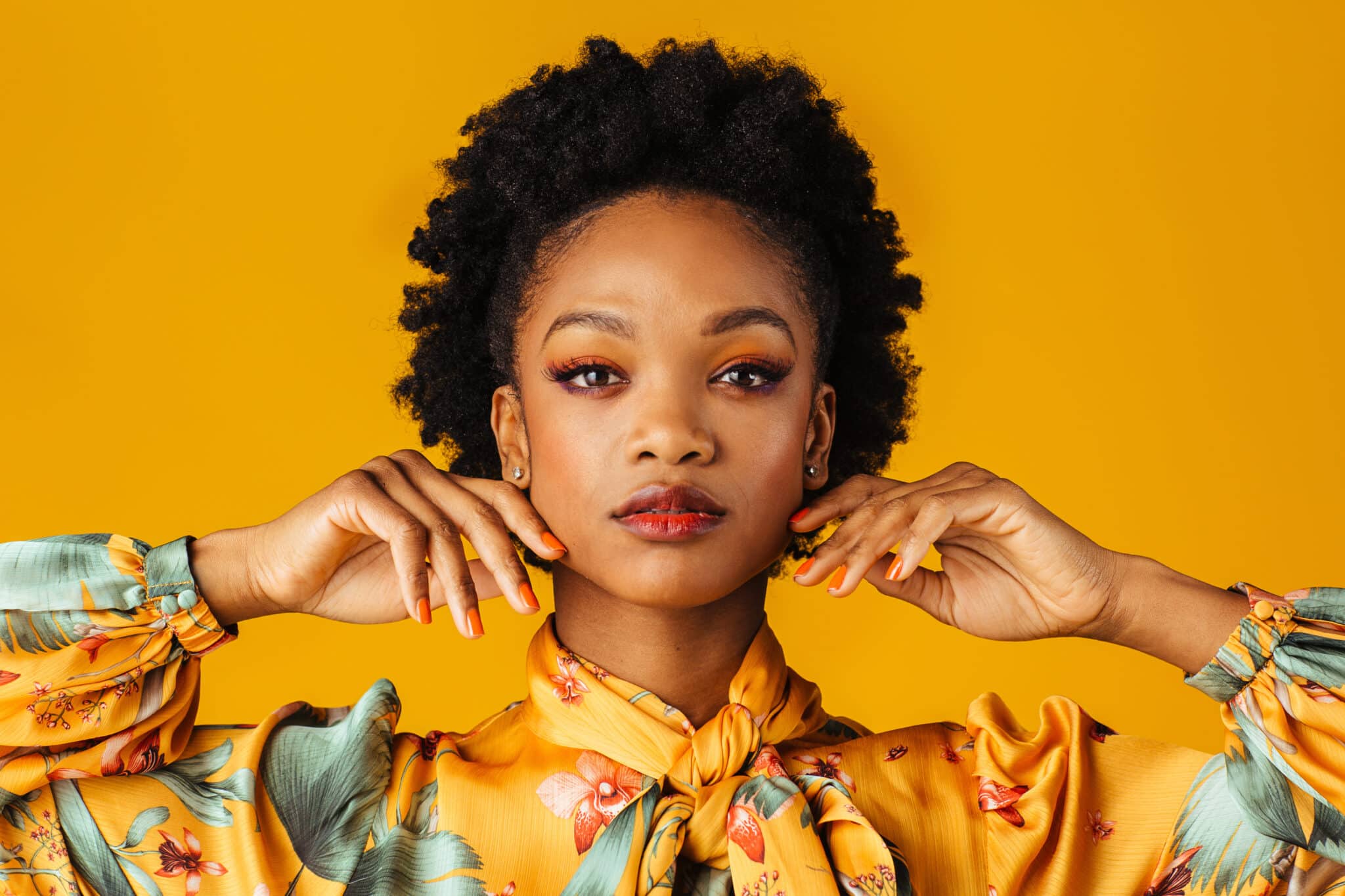 A woman with fresh hair poses confidently against a vibrant yellow background. She is wearing a yellow floral blouse and has her hands gently touching her neck, showcasing orange nails and matching eye makeup. She gazes directly at the camera with a calm expression that reflects her dedication to organic hair care.