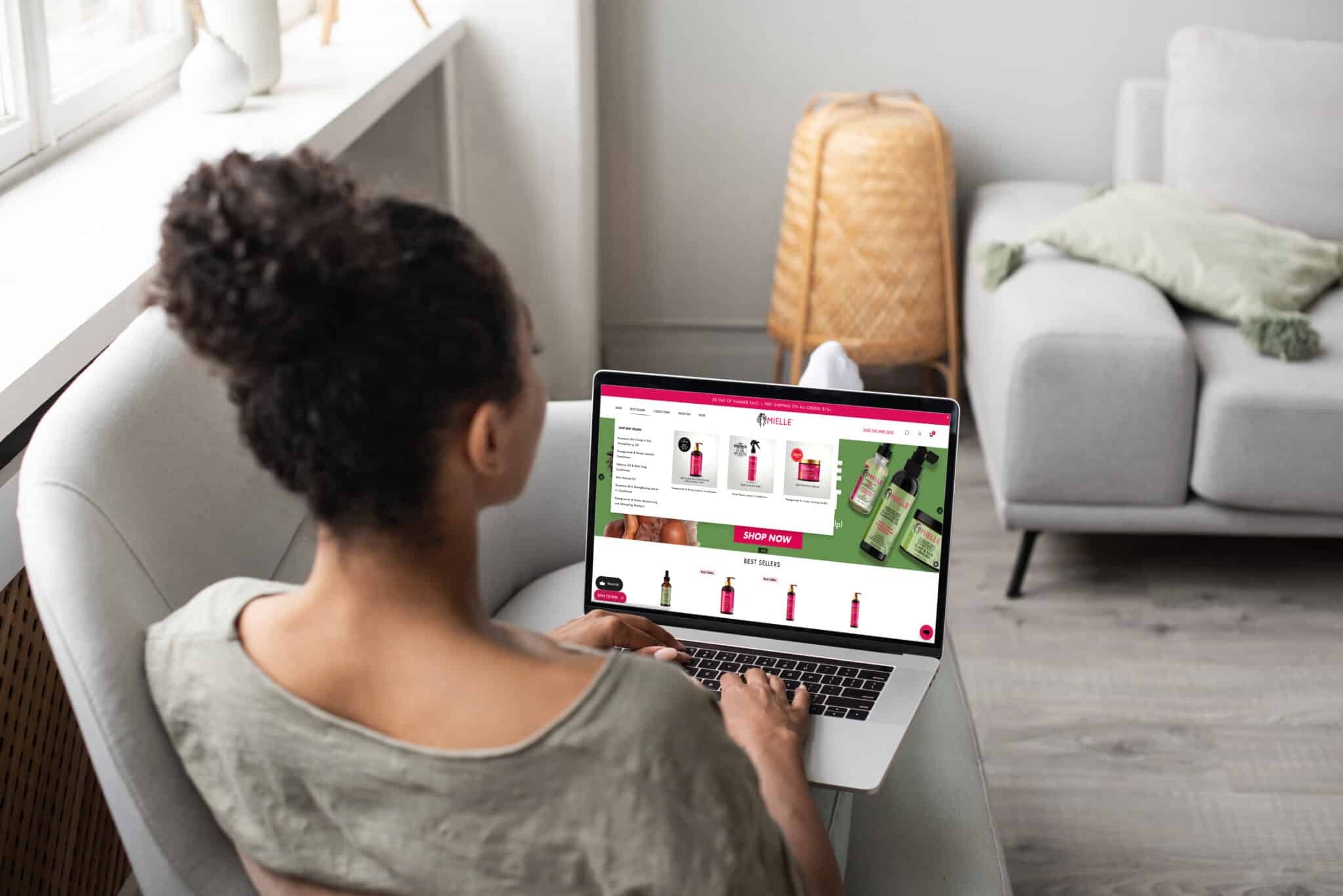 A person with curly hair in a bun is sitting on a white chair, using a laptop, browsing an online shopping website featuring various skincare products from Mielle Organics. The background includes a beige rug, a wicker basket, a light green blanket, and a gray sofa.