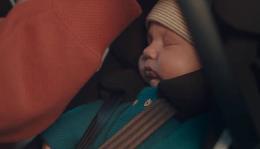 A baby wearing a striped beanie and teal top is peacefully sleeping in a Bugaboo car seat. The baby is secured with a safety belt. A person's arm, dressed in a red sleeve, is gently touching the baby.