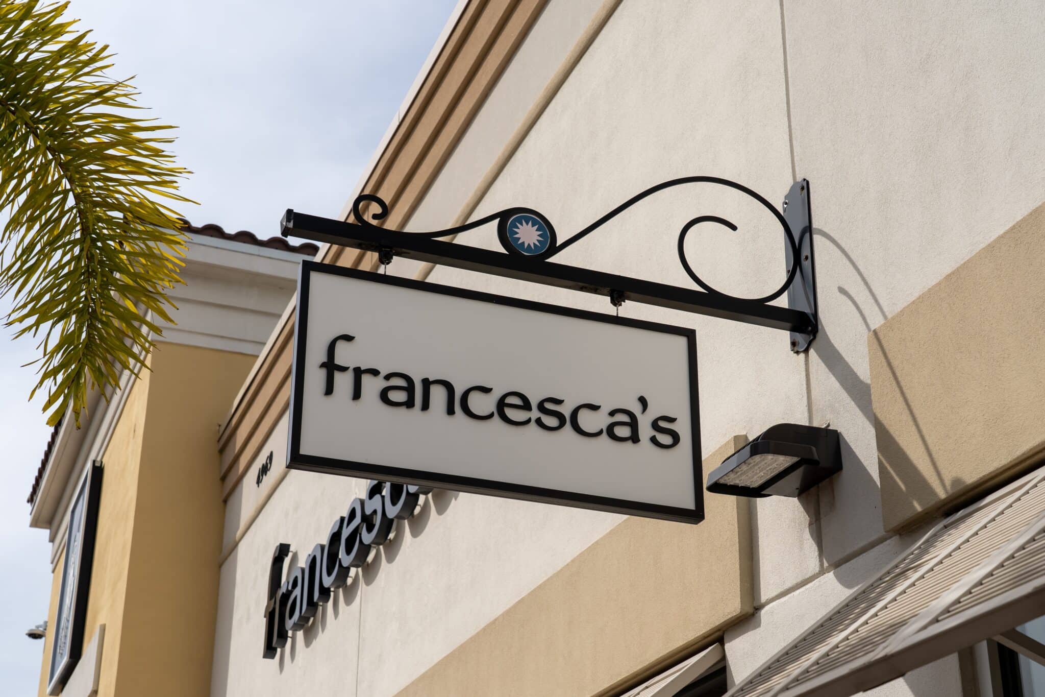 Storefront of Francesca's with a hanging sign featuring the store's name. The sign, embodying a blend of simple and modern design, is attached to a cream-colored wall. Part of a palm tree is visible on the left side, capturing Francesca's transformation from brick-and-mortar to an eCommerce-savvy brand.