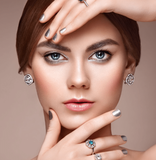 A woman with bright blue eyes gazes forward, her hands framing her face. She has light pink blush, metallic nail polish, and is adorned with several pieces of jewelry, including gem-studded rings, earrings, and a hand accessory—appearing every bit the poised media executive.
