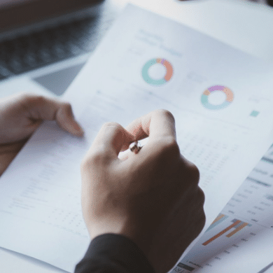 A person holding a pen reviews a financial document. The paper features charts, graphs, and tables with colorful circular graphs visible near the top. In the background, a keyboard and another sheet of paper indicate the focus on marketing analytics in B2B sales.