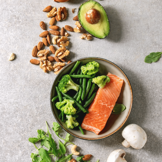 A healthy meal featuring raw salmon, green beans, and broccoli in a bowl is presented for your next video ad. Scattered around the bowl are almonds, walnuts, a halved avocado, mushrooms, and arugula leaves on a gray surface.