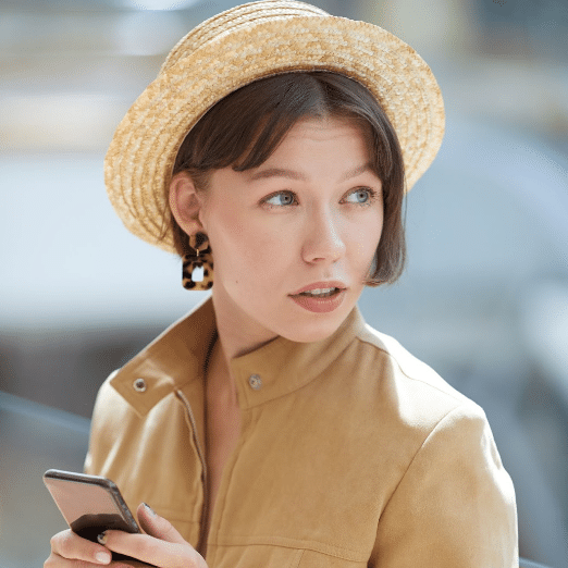 A young woman with short brown hair, wearing a straw hat and leopard print earrings, gazes to the side while holding a smartphone. She has a neutral expression and is dressed in a light brown jacket, exuding an air of professionalism. The softly blurred background hints at her involvement in global PR.
