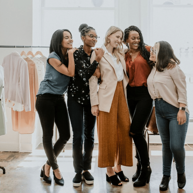 A group of five women, standing close together and smiling, in a brightly lit room with clothing racks and hangers in the background. Sporting casual yet stylish outfits, they reflect a friendly and joyful atmosphere perfect for a Paid Social campaign.