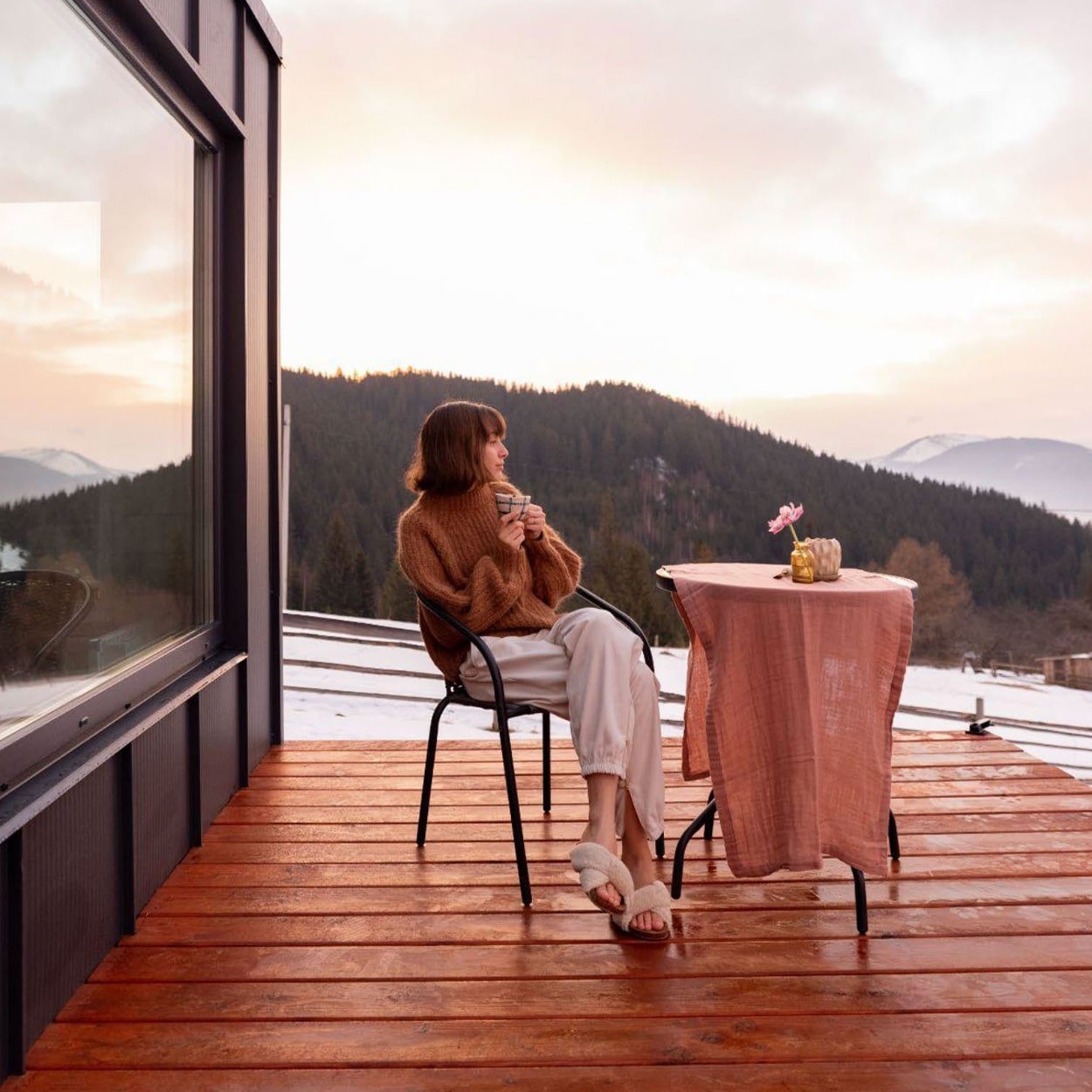 A person sits on a chair on a wooden terrace, wearing a brown sweater and white pants, holding a mug. The terrace overlooks a scenic mountain landscape at sunset. A small table with a pink tablecloth and a vase with a flower is beside them.