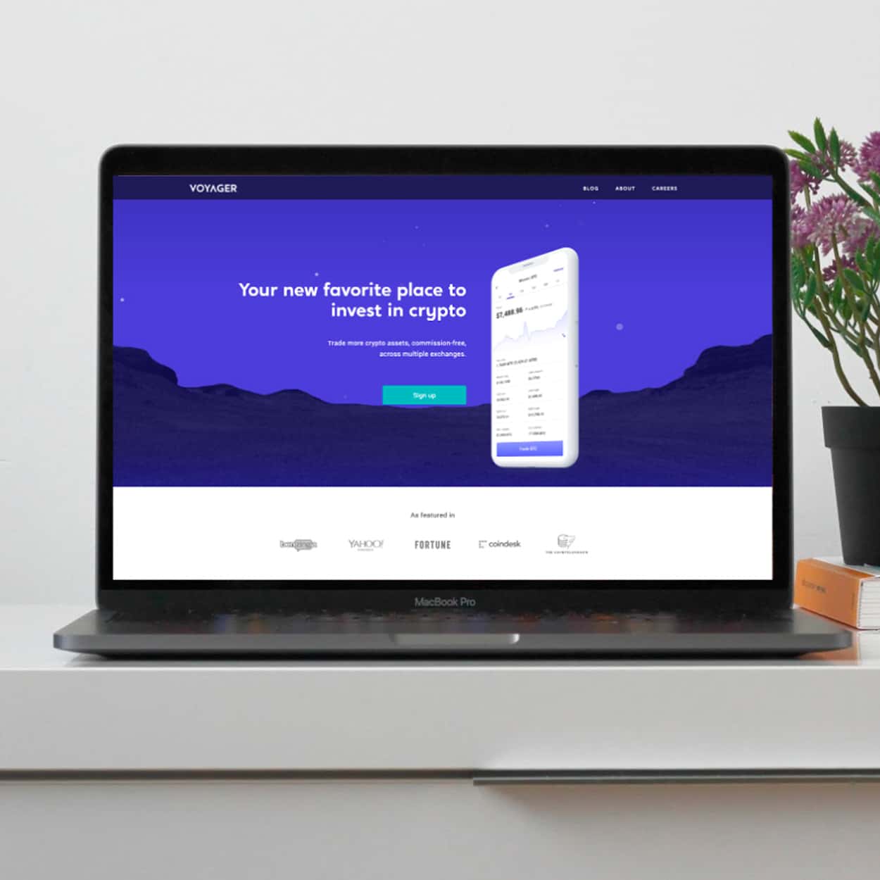 A laptop displaying Voyager's cryptocurrency investment platform with a blue and white interface. The screen shows the text, "Your new favorite place to invest in crypto," above an image of their mobile app. The laptop is on a white desk beside a potted plant, ensuring an optimal CX.