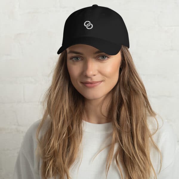 A woman with long, wavy hair is wearing a black Dad Hat with a circular logo on the front. She is dressed in a white sweatshirt and is standing against a plain, light-colored brick wall. She has a slight smile and appears relaxed.