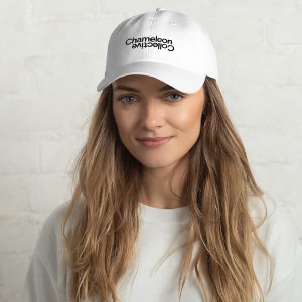 A woman with long, blonde hair smiles softly while sporting a Dad hat with the text "Chameleon Collective" embroidered in black. She is dressed in a casual white top, and the background is a plain, white brick wall.
