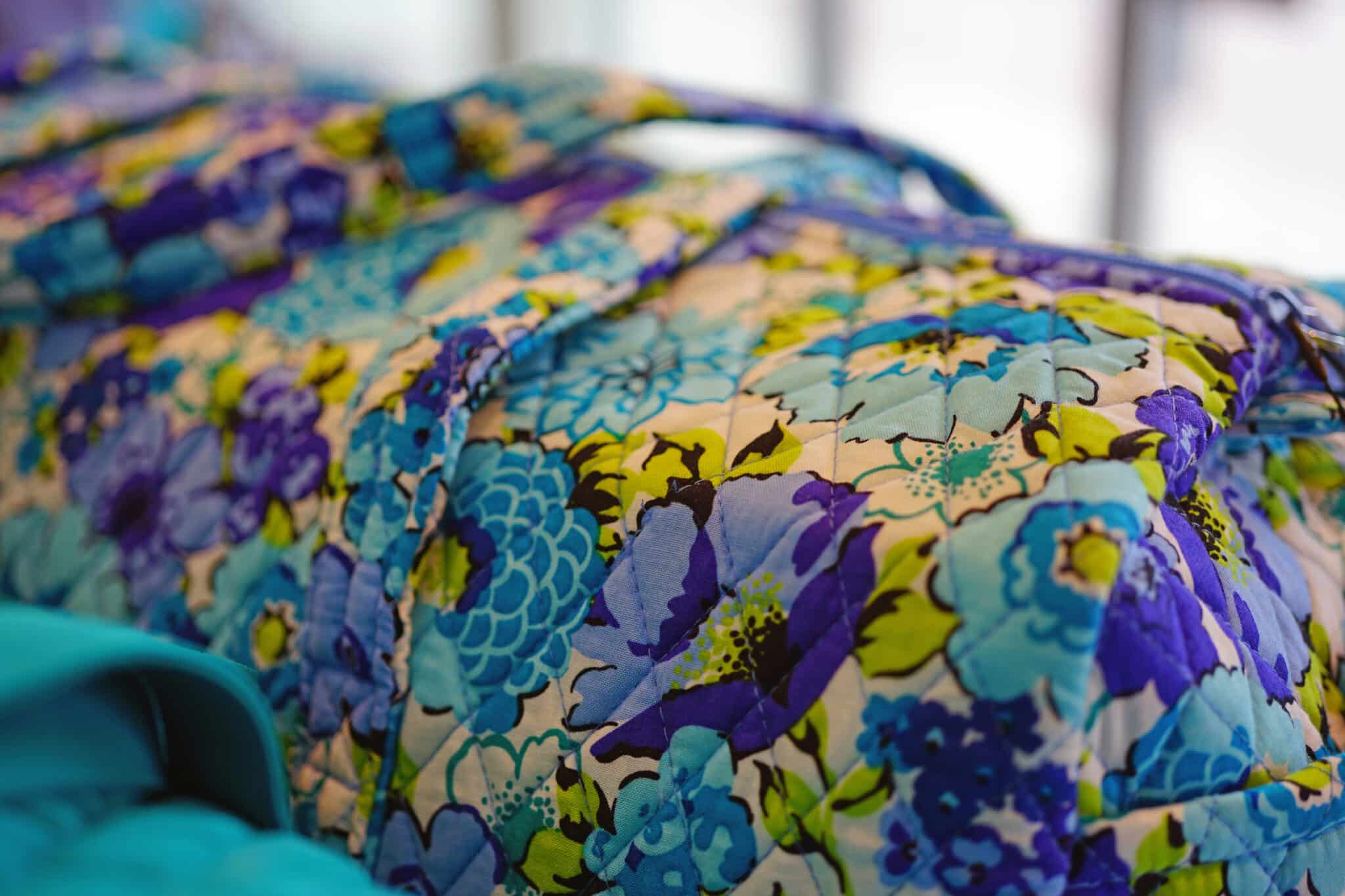 A close-up view of a fabric with a vibrant floral pattern in shades of blue, purple, and green. The print features large flowers on a textured surface, suggesting the item could be an iconic Vera Bradley bag or piece of clothing. Bright lighting enhances the vivid colors.