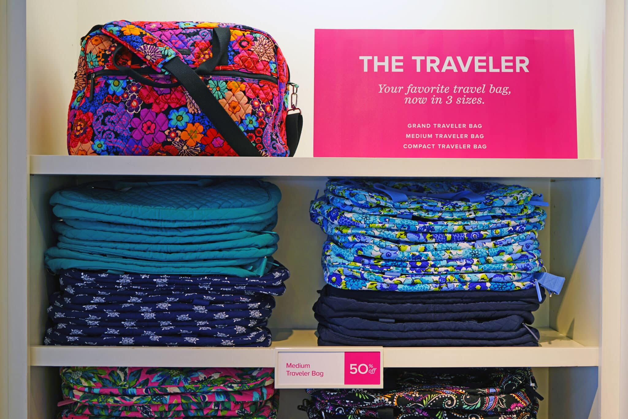 Shelves displaying various colorful bags. The top shelf features a floral-patterned travel bag, next to a pink sign reading "The Traveler: Your favorite travel bag, now in 3 sizes." Iconic Vera Bradley folded travel bags fill the lower shelves, accompanied by a sign reading "Medium Traveler Bag - 50% Off.