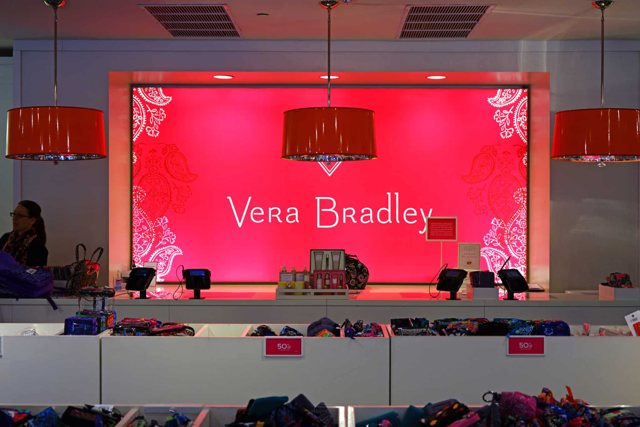 A brightly lit Vera Bradley store display features a large pink backdrop with the brand name centered in white text. Red pendant lights hang above the counter below, which has bags and accessories neatly arranged, and a sign indicating a sale. Truly, always iconic!