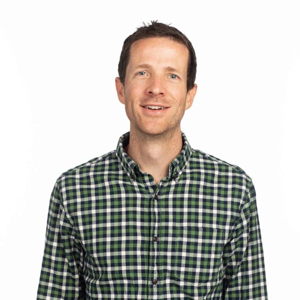 Ben Barlow, a man with short brown hair, is smiling while wearing a green and blue plaid shirt. He is standing against a plain white background.