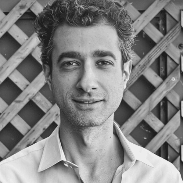Black and white photo of a man with curly hair, identified as Ilan Tito, wearing a collared shirt. He is standing in front of a wooden lattice backdrop and looking at the camera with a neutral expression.