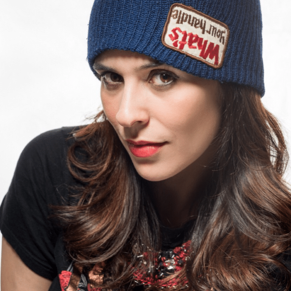 A person with long dark hair wearing a blue beanie with a patch that reads "Max's Kansas City" and a black shirt. Ivonne Azurdia is looking directly at the camera with a neutral expression, and the background is plain white.
