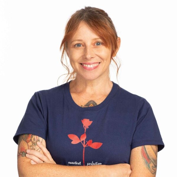 Michelle Barry, a woman with red hair pulled back, smiles confidently with her arms crossed. She is wearing a navy blue t-shirt featuring a red flower design. Tattoos are visible on both arms, and her background is plain white.