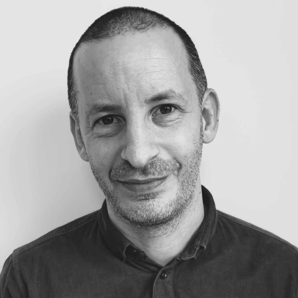 A man with a shaved head and light beard is looking at the camera. He is wearing a dark button-up shirt and is set against a plain white background. This striking portrait captures Rob Gotlieb's confident gaze and distinctive style.