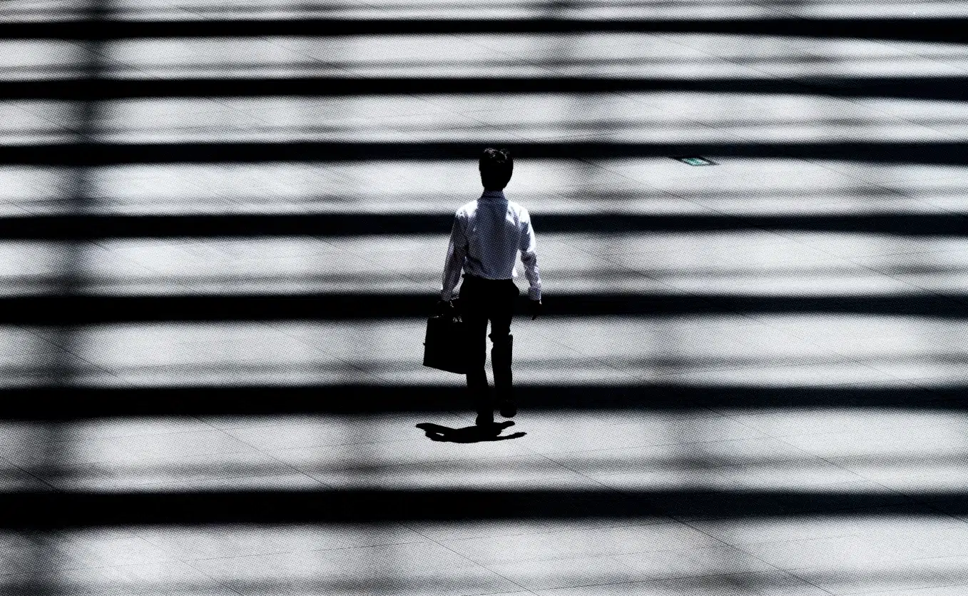 A person in business attire is walking alone on a sunlit street, casting a long shadow. The ground is patterned with contrasting light and dark stripes, creating bold shadows that add a dramatic effect to the scene. With a briefcase in their right hand, they seem intent on heading home.
