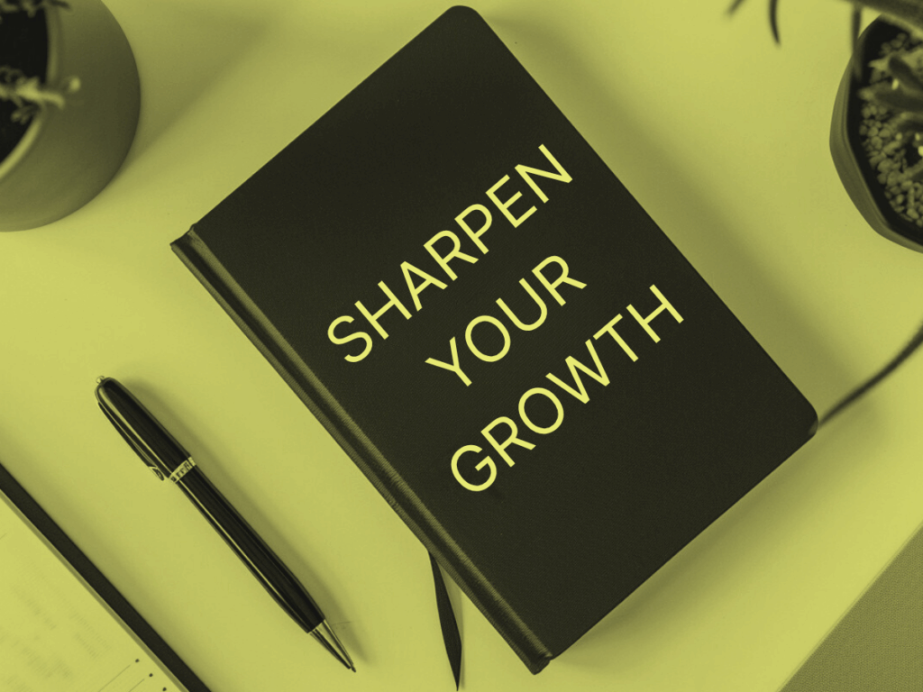 Black hardcover notebook with white text reading "Sharpen Your Growth," placed on a light desk with a pen, notepad, and small plants