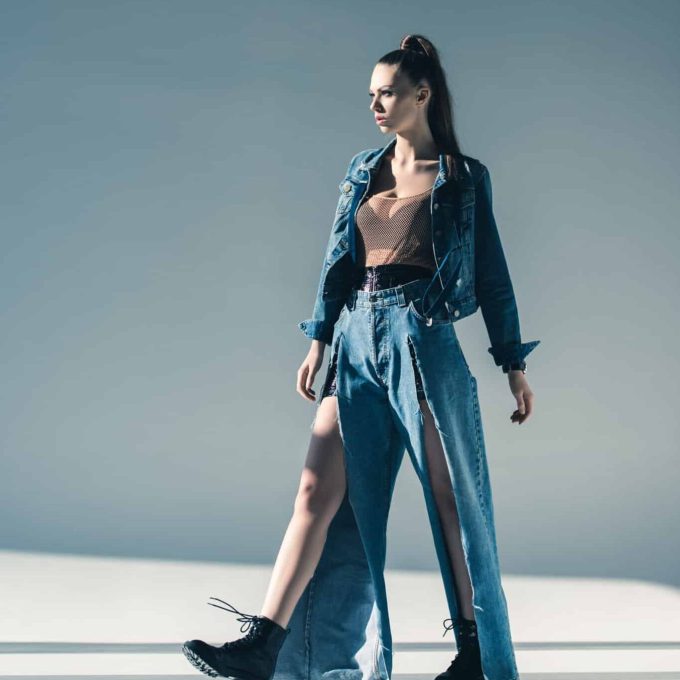 A woman stands confidently in a fashionable outfit against a minimalist background. She wears a denim jacket, a sheer brown top, and unique wide-leg jeans with high side slits. Her hair is tied in a high ponytail, and she pairs the look with black combat boots, exuding an air of international sophistication.