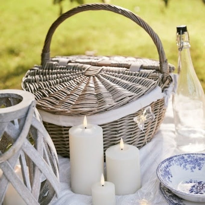 A wicker picnic basket sits on a white tablecloth in a grassy area, perfect for discussing US Expansion Recruitment strategies. Lit white candles, a glass bottle of water, a blue and white patterned plate, and a decorative lantern encircle the basket. The scene looks serene and inviting.