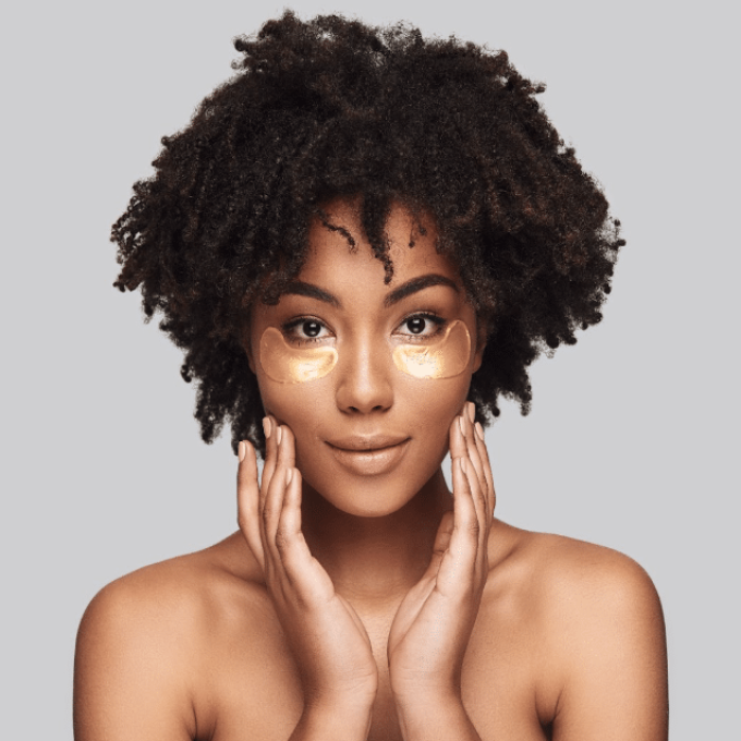 A person with curly hair is posing against a plain background, with under-eye patches applied beneath their eyes—a testament to their dedicated skincare routine. Their hands are gently resting on their cheeks as they look directly at the camera with the calm confidence of a creative leader.