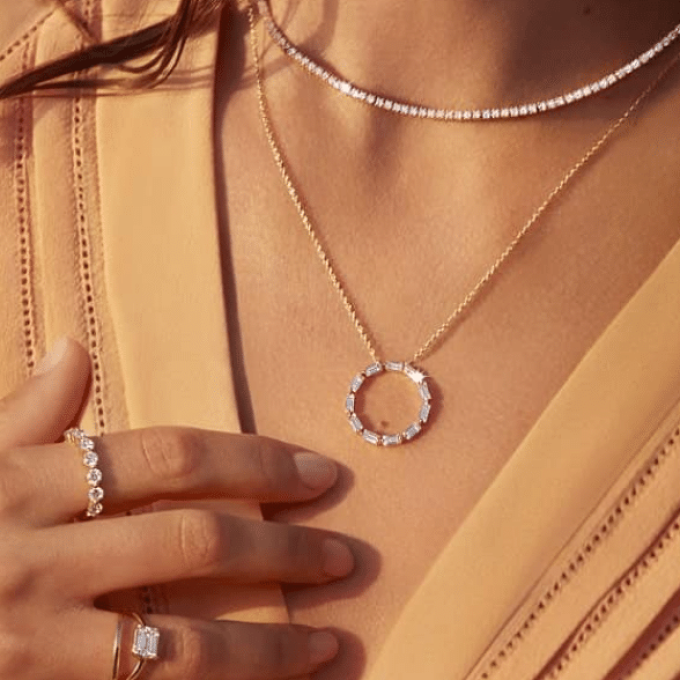 A person wearing a light peach dress is shown from the neck to the chest, exuding luxury with several pieces of jewelry. They are wearing a thin necklace with small gems, a gold necklace with a circular gem-studded pendant, and rings on their fingers, including one with a rectangular gem.