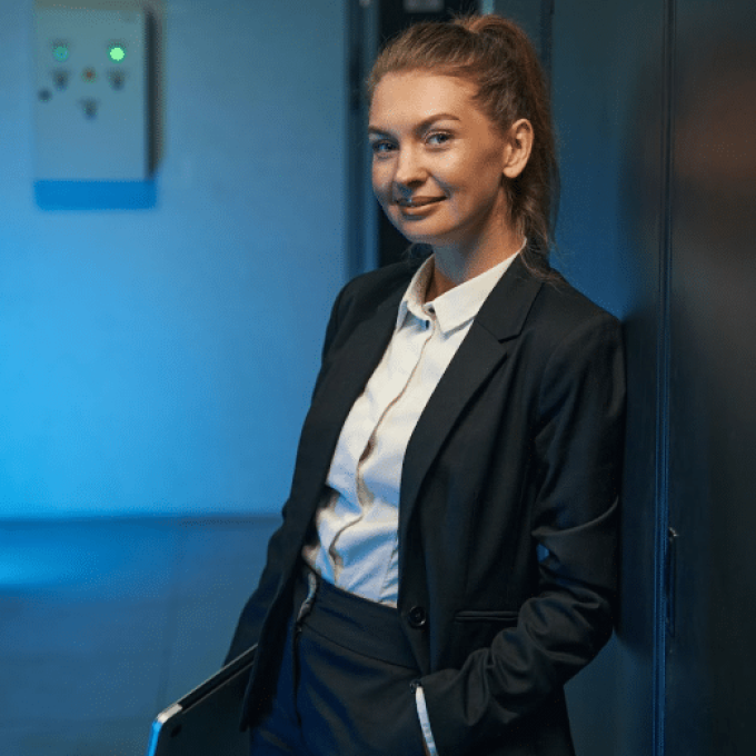 A woman with long brown hair tied in a ponytail, wearing a dark business suit and white shirt, smiles softly while standing against a wall with her hand in her pocket. She holds a closed laptop under her arm, ready for the recruiting meeting. The background is lit with blue light.