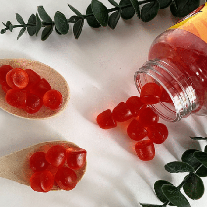 A bottle of red gummy vitamins tips over, spilling some gummies onto a white surface. Two wooden spoons, each holding a few red gummies, are placed nearby. Green leafy branches are arranged decoratively around the scene, highlighting the Direct-to-Consumer appeal.
