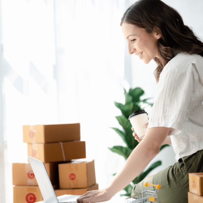 A woman is smiling, holding a coffee cup, and working on a laptop at a desk. Several cardboard boxes are stacked in the background. The room is bright with sunlight streaming through a window, and a plant is visible in the background, as she focuses on her recruitment tasks for her team.