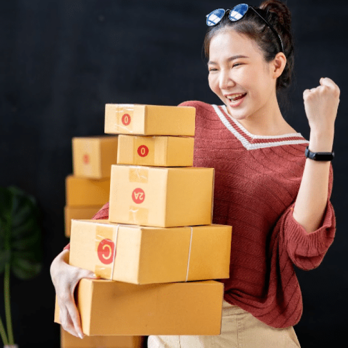 A woman wearing a red sweater and sunglasses on her head is smiling and holding a stack of cardboard boxes. She is indoors with more boxes in the background, appearing excited and raising her left fist in a celebratory gesture, embodying the spirit of Business Development.