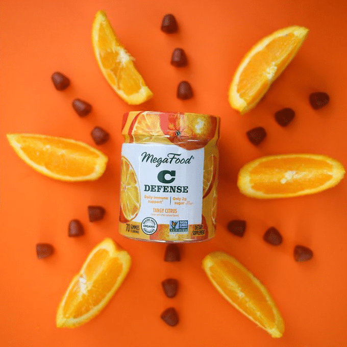 A container of MegaFood Vitamin C Defense gummies is surrounded by orange slices and individual gummies, creating a visually appealing pattern on an orange background, perfect for eCommerce displays.