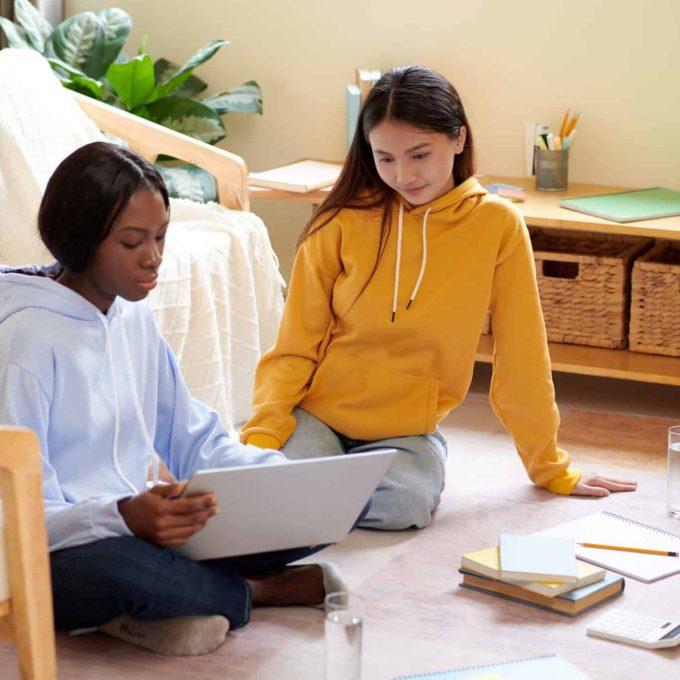 Two young women, dressed in casual hoodies, study together on the floor of a cozy, well-lit room. One woman writes in a notebook while the other observes. Books, papers, and stationery are scattered around, creating an atmosphere of strategic learning and leadership development.